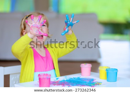 Laughing little child, blonde artistic toddler girl painting and drawing with colorful finger paints indoors at bright room at home or kindergarten. Focus on messy hands