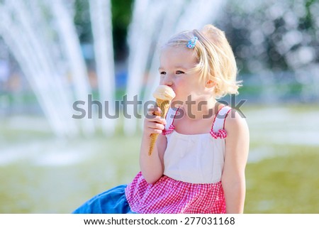Summer in town. Happy child enjoying hot day outdoors. Cute little toddler girl eating ice cream outdoors in city park. Beautiful blurred fountain at background.