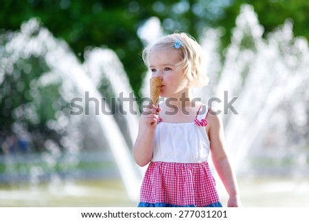 Summer in town. Happy child enjoying hot day outdoors. Cute little toddler girl eating ice cream outdoors in city park. Beautiful blurred fountain at background.