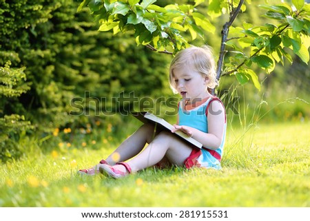 Happy preschooler child reading interesting book in the garden. Cute little girl relaxing and playing outdoors in the park enjoying sunny summer day.
