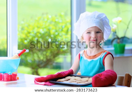 Little happy child, adorable toddler girl wearing white chef hat and red gloves helping mother cooking delicious pastry in the kitchen