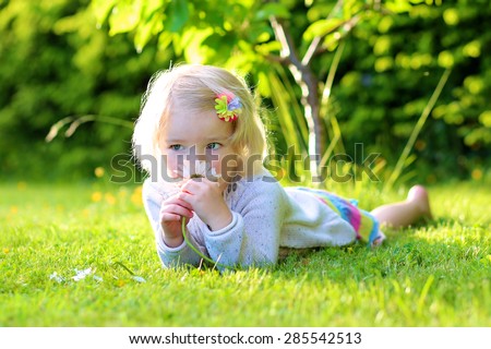 Little happy child, smiling blonde toddler girl lying on green grass holding daisy flowers playing he loves me or not