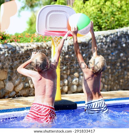 Two happy teenage boys, sportive twin brothers, having fun together playing basketball with colorful inflatable ball in outdoors swimming pool in aquapark during summer sea vacation in tropical resort