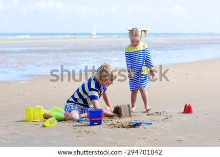 Two happy active children, teenage boy with his little sister, cute blonde toddler girl, playing with plastic toys building sand castles sitting on wide sandy North European beach