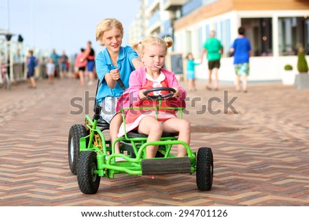 Happy kids enjoying active holidays on the beach. Sportive boy riding pedal car together with his cute toddler sister along the promenade on a summer day at sunset.