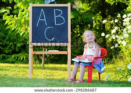 Little preschooler girl excited to go back to school. Cute toddler playing teacher role game outdoors. Happy kid reading book, leaning letters and numbers at kindergarten. Children education concept.