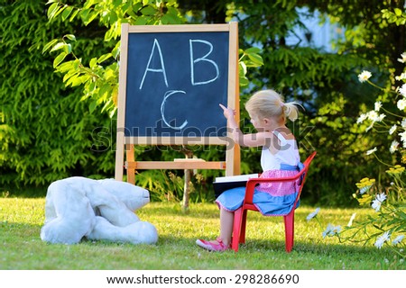 Little preschooler girl excited to go back to school. Cute toddler playing teacher role game outdoors. Happy kid leaning letters and number at kindergarten. Children education concept.