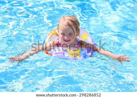 Adorable happy little child, curly toddler girl in swimming suit having fun relaxing and floating on an inflatable toy ring in a pool on sunny day during summer vacation in resort