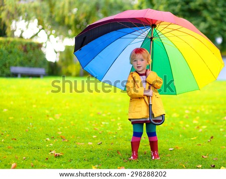Happy little child, adorable blonde curly toddler girl wearing yellow waterproof coat and red boots holding colorful umbrella playing in the garden or park on a sunny rainy warm early autumn day