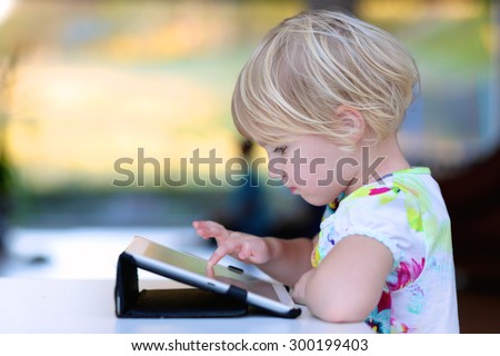 Happy little child, adorable blonde toddler girl enjoying modern generation technologies playing indoors using tablet pc with touchscreen