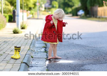 Happy little child, adorable blonde curly toddler girl wearing red duffle coat enjoying sun after rain running barefoot and jumping on the puddle on the street on a sunny autumn or spring day