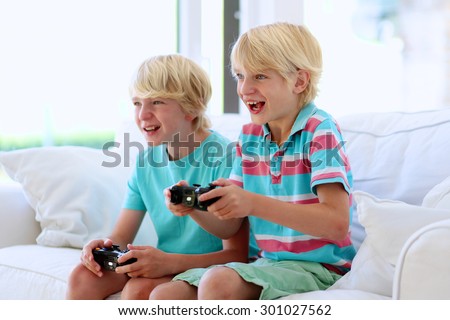 Two kids playing video game at home. Twin teenage brothers having fun after school day laughing and holding joysticks in hands sitting on white sofa in bright sunny living room with big windows
