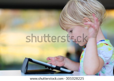Happy little child, adorable blonde toddler girl enjoying modern generation technologies playing indoors using tablet pc with touchscreen.