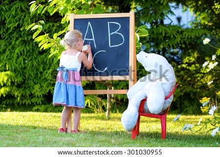 Little preschooler girl excited to go back to school. Cute toddler playing teacher role game outdoors. Happy kid leaning letters at kindergarten. Children education concept.