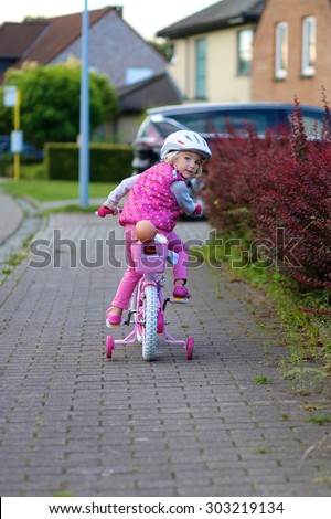 Little child riding pink bicycle on the street in countryside. Cute preschooler girl learning to cycle with stabilisers wheels. Sportive kid enjoying sunny autumn day outdoors.