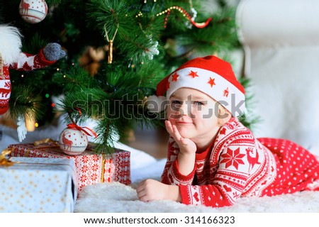 Little smiling girl wearing Santa hat and festive dress lying on the lambskin carpet near the Christmas tree and gifts. Adorable kid making new year wishes.