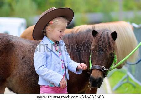 Lovely cowgirl caressing little pony horse in the farm. Pretty preschooler girl wearing cowboy hat playing with animals outdoors on sunny day.