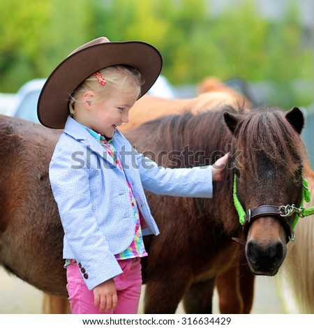 Lovely cowgirl caressing little pony horse in the farm. Pretty preschooler girl wearing cowboy hat playing with animals outdoors on sunny day.