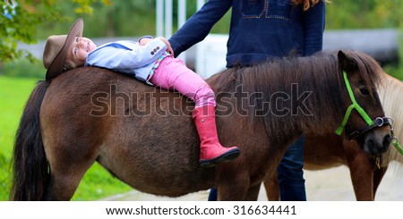 Lovely cowgirl lying on the back of little pony horse in the farm. Pretty preschooler girl wearing cowboy hat playing with animals outdoors on sunny day.