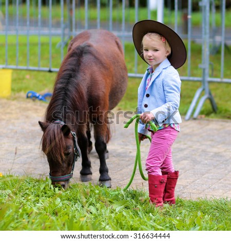 Lovely cowgirl feeding with grass little pony horse in the farm. Pretty preschooler girl wearing cowboy hat playing with animals outdoors on sunny day.