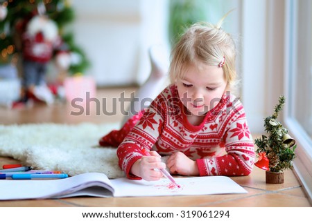 Cute little kid  writes letter to Santa. Adorable toddler girl in festive dress lying on the floor on lambskin carpet. Christmas tree with lights and gifts at the background.