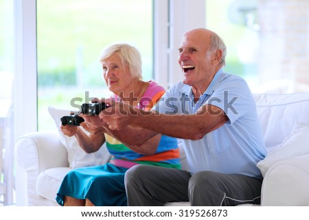 Senior couple having fun at home playing video game holding joysticks in hands sitting cozy on the sofa in bright sunny living room with big windows. Happy retirement concept.