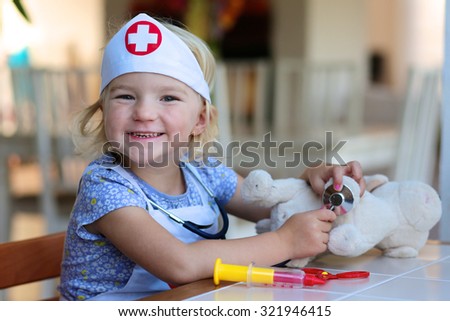 Funny blonde toddler girl playing doctor role game examinating her puppy using stethoscope sitting at small white table in playroom at home, school or kindergarten