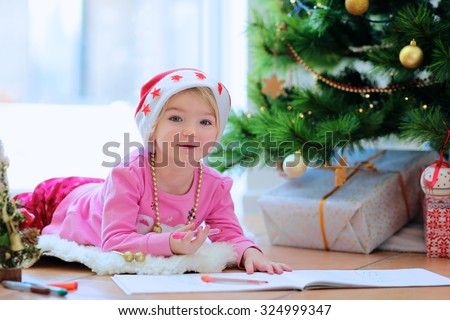 Cute little kid writes letter to Santa. Adorable toddler girl in Santa hat lying on the floor on lambskin carpet next to big window. Christmas tree with lights and gifts at the background.