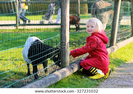 Adorable toddler girl petting little goat in the kids farm. Cute kind child feeding animals in the zoo. Family with preschooler enjoying day in the park.