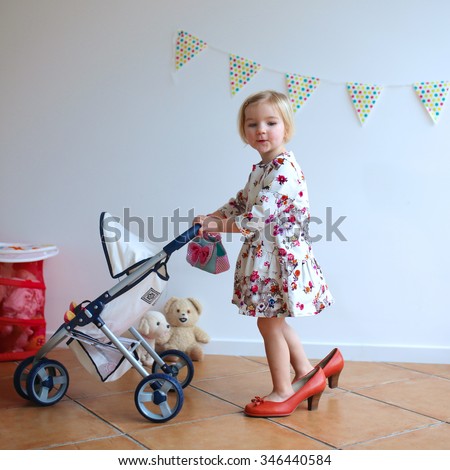 Happy little child, cute blonde toddler girl, wearing beautiful dress and red mom's shoes playing role game pushing stroller with baby doll indoors at home or kindergarten