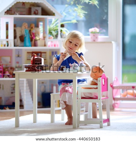 Cute preschooler girl having birthday tea party with her doll. Little child plays in sunny room at home or kindergarten. Kids using toy dishes and cakes.
