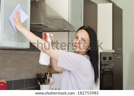 Smiling dark haired woman washing the furniture in the kitchen with cloth and spray cleaner, looking at camera