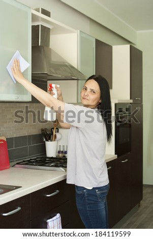 Beautiful young woman in jeans cleaning furniture in kitchen, looking at camera
