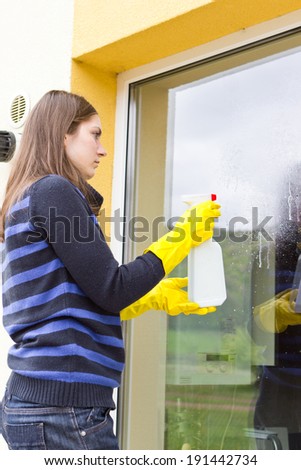 Young female maid in yellow gloves washing facade windows with spray cleaner