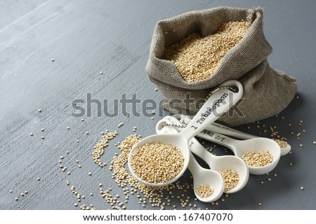 Quinoa grain in small burlap sack and porcelain measuring spoons on gray background