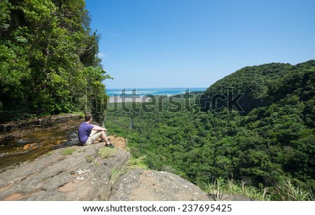 Man sitting on cliff overlooking beautiful view of tropical jungle and turquoise ocean in Okinawa, Japan