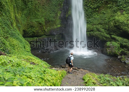Hiker standing in front of huge Jungle waterfall surrounded by lush Jungle vegetation and flora in Lombok, Indonesia