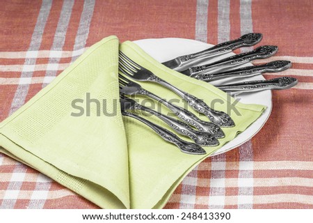 A white plate with a green napkin and set of forks and knifes on a red tablecloth