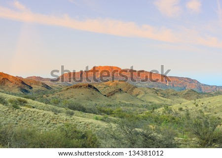 Green mountain landscape with in the background the glowing red mountain cliff of the Gammon Ranges, South Australia, against a blue sky with clouds veil