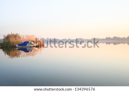 A lonely little boat is moored near a Reed border and reflected on the water surface in the Orange morning sun on a smoothly lake against a misty horizon.