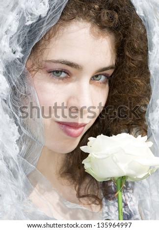 Attractive young woman with a bridal veil is smiling at the camera and smelling a white rose. Wedding and celebration concept.