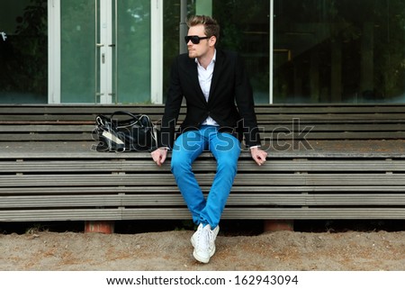 A man sitting outside wearing black sunglasses and a black jacket with blue jeans.