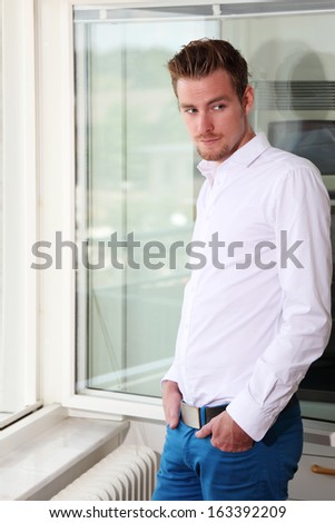 Good looking young man standing in front of a window looking out. Wearing a white shirt and blue jeans.