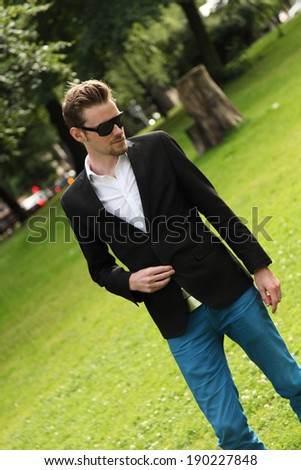 A young attractive man standing on green grass wearing a black jacket and black sunglasses.