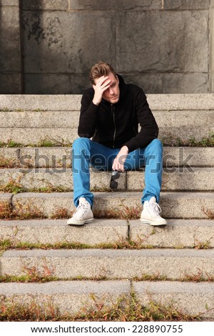 A young man wearing blue jeans and a black hoodie sitting down outside with sunglasses, sitting down on steps of stone.