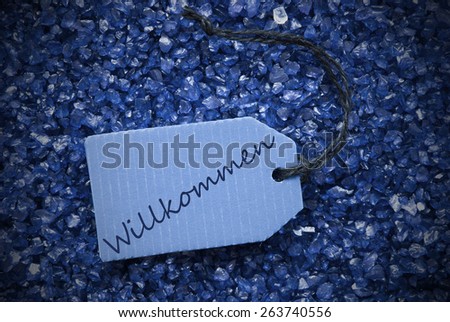 One Blue Label Or Tag With Black Ribbon On Blue And Purple Small Stones As Background With German Text Willkommen Means Welcome With Frame