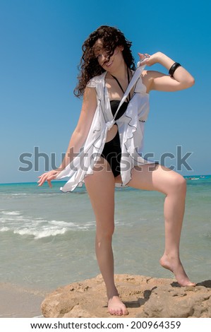 Girl in a black bathing suit with a white cape dancing on a cliff above the sea