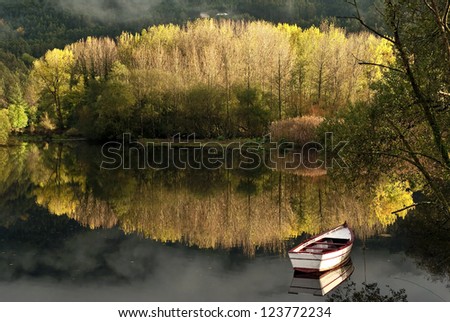 A nearby forest fall colors are reflected in the calm of a lake where there is a boat./Autumn reflection