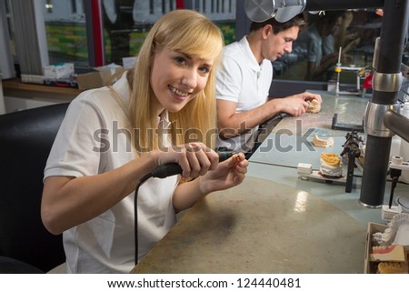 Female dental technician polishing a gold tooth in a dental lab. Another technician producing a dental prosthesis in the background