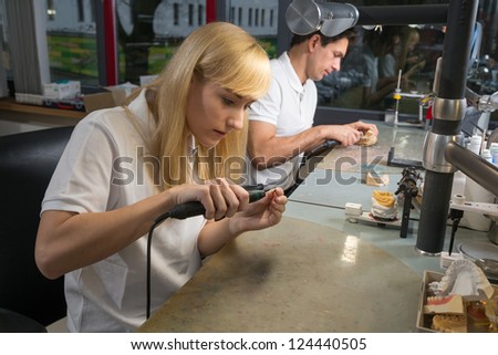Female dental technician polishing a gold tooth in a dental lab. Another technician producing a dental prosthesis in the background